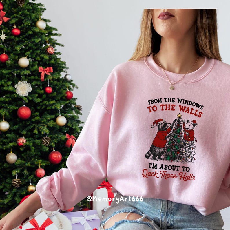 May include: A light pink sweatshirt with the text "From the Windows to the Walls, I'm About to Deck These Halls." The graphic features two raccoons decorating a Christmas tree. A Christmas tree is visible in the background.