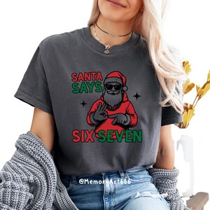 May include: Dark gray t-shirt with a graphic of Santa Claus wearing sunglasses and a red hoodie. The text "SANTA SAYS SIX SEVEN" is printed in green and red. The shirt is a casual style.