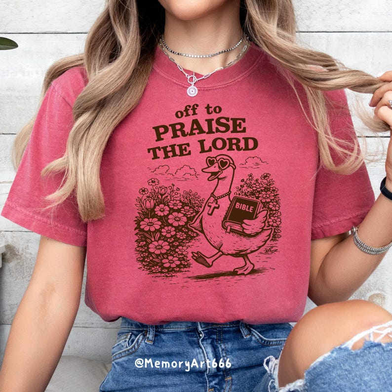 Op de afbeelding: Een bordeauxrood T-shirt met de tekst "off to PRAISE THE LORD" boven een cartoon gans met een hartvormige zonnebril en een bijbel. De gans loopt door een bloementuin. Het shirt heeft een effen kleur.
