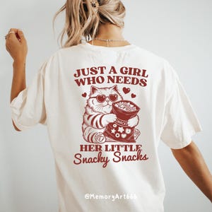May include: White t-shirt with a red graphic of a cat holding a bag of snacks. The text on the shirt reads "Just a girl who needs her little snacky snacks" and "@MemoryArt666".