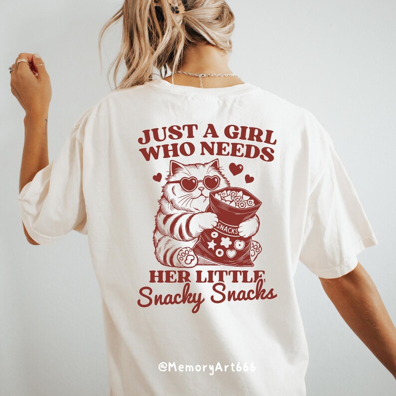 May include: White t-shirt with a red graphic of a cat holding a bag of snacks. The text on the shirt reads "Just a girl who needs her little snacky snacks" and "@MemoryArt666".
