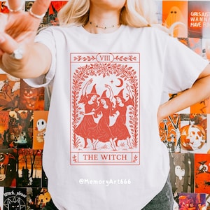 May include: White t-shirt featuring a red tarot card design with three witches dancing under a crescent moon and stars. The card reads "THE WITCH" and "VIII". The shirt has the Instagram handle @MemoryArt666.