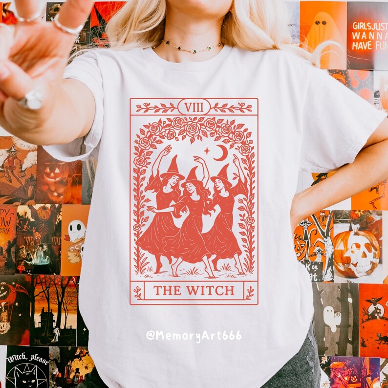 May include: White t-shirt featuring a red tarot card design with three witches dancing under a crescent moon and stars. The card reads "THE WITCH" and "VIII". The shirt has the Instagram handle @MemoryArt666.