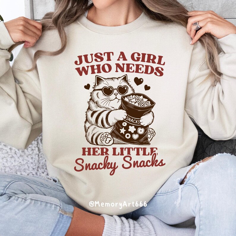 May include: A beige sweatshirt with the text "Just a girl who needs her little snacky snacks" and a cartoon cat wearing sunglasses holding a bag of snacks. The text is in brown and red. The cat is brown and white. The bag of snacks is brown and white with the word "snacks" printed on it.