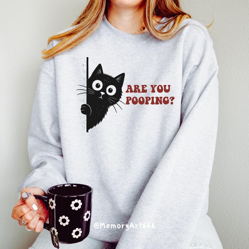 May include: A light gray sweatshirt with a black cat graphic peeking from behind a black line. The text "ARE YOU POOPING?" is printed in maroon. A black mug with white flower designs is held in the hand.