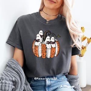 May include: Dark gray t-shirt featuring a Halloween-themed design. The graphic depicts a pumpkin made of books with ghosts reading inside. The design includes orange, white, brown, and green colors. The shirt has short sleeves.