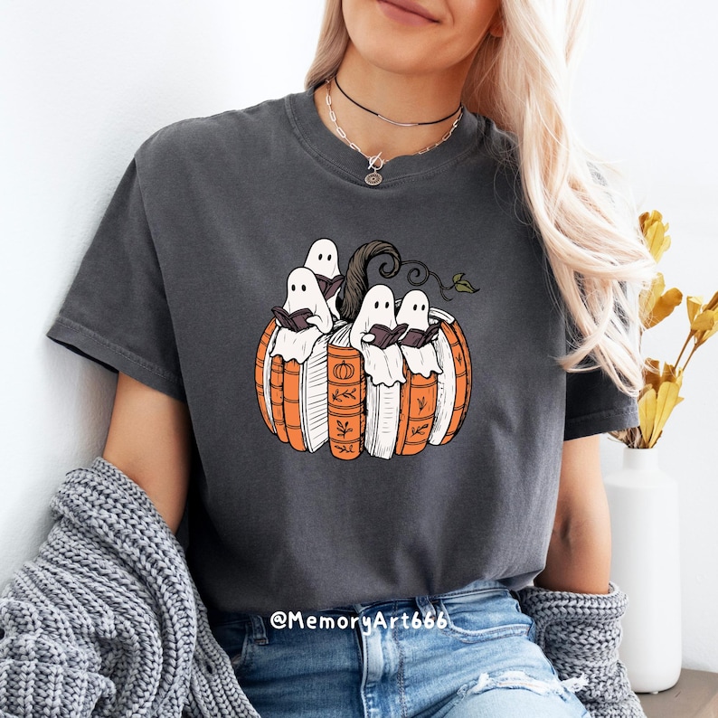 May include: Dark gray t-shirt featuring a Halloween-themed design. The graphic depicts a pumpkin made of books with ghosts reading inside. The design includes orange, white, brown, and green colors. The shirt has short sleeves.