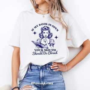 May include: White t-shirt with a blue graphic of a woman reading a book and saying "If my book is open your mouth should be closed".