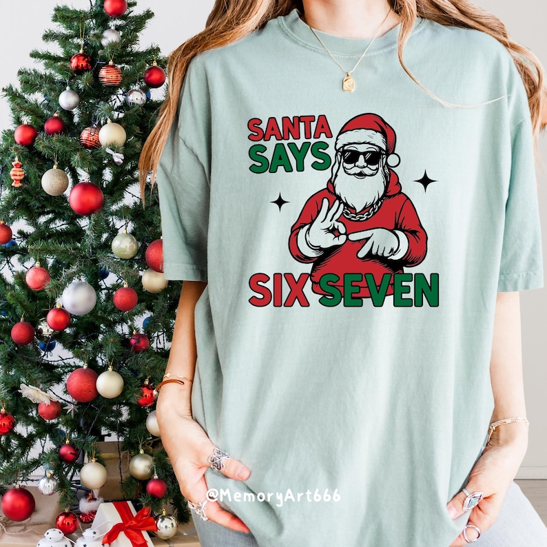 May include: A light blue t-shirt with a graphic of Santa Claus wearing sunglasses and a red outfit. The text "SANTA SAYS SIX SEVEN" is printed in red and green. A Christmas tree is in the background.
