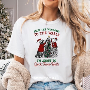 May include: White t-shirt with a Christmas-themed graphic. The design features two cartoon animals, a raccoon and a mouse, decorating a Christmas tree. Text on the shirt reads "From the windows to the walls, I'm about to Deck These Halls."