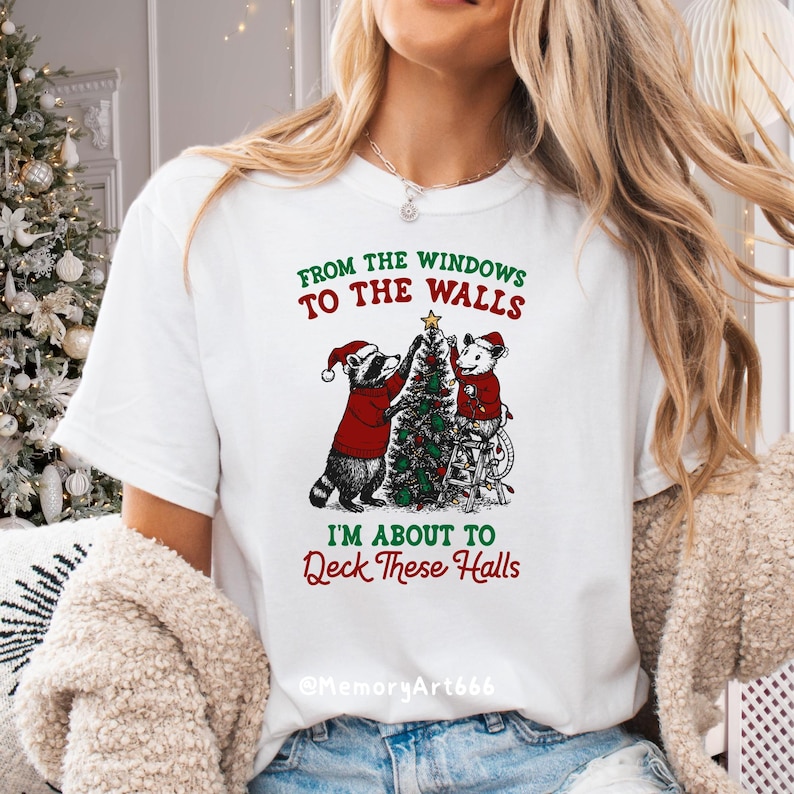 May include: White t-shirt with a Christmas-themed graphic. The design features two cartoon animals, a raccoon and a mouse, decorating a Christmas tree. Text on the shirt reads "From the windows to the walls, I'm about to Deck These Halls."