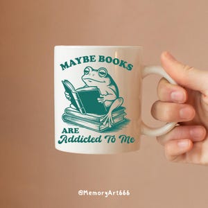 May include: White ceramic mug with a teal frog reading a book, with the text "MAYBE BOOKS ARE ADDICTED TO ME". The mug has a curved handle and is held by a hand.