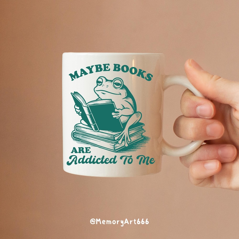 May include: White ceramic mug with a teal frog reading a book, with the text "MAYBE BOOKS ARE ADDICTED TO ME". The mug has a curved handle and is held by a hand.