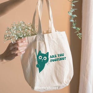 May include: Cream-colored tote bag with a teal cat graphic and the text "ARE YOU POOPING?". The bag has long handles and is filled with white flowers. The bag is made of a sturdy fabric.