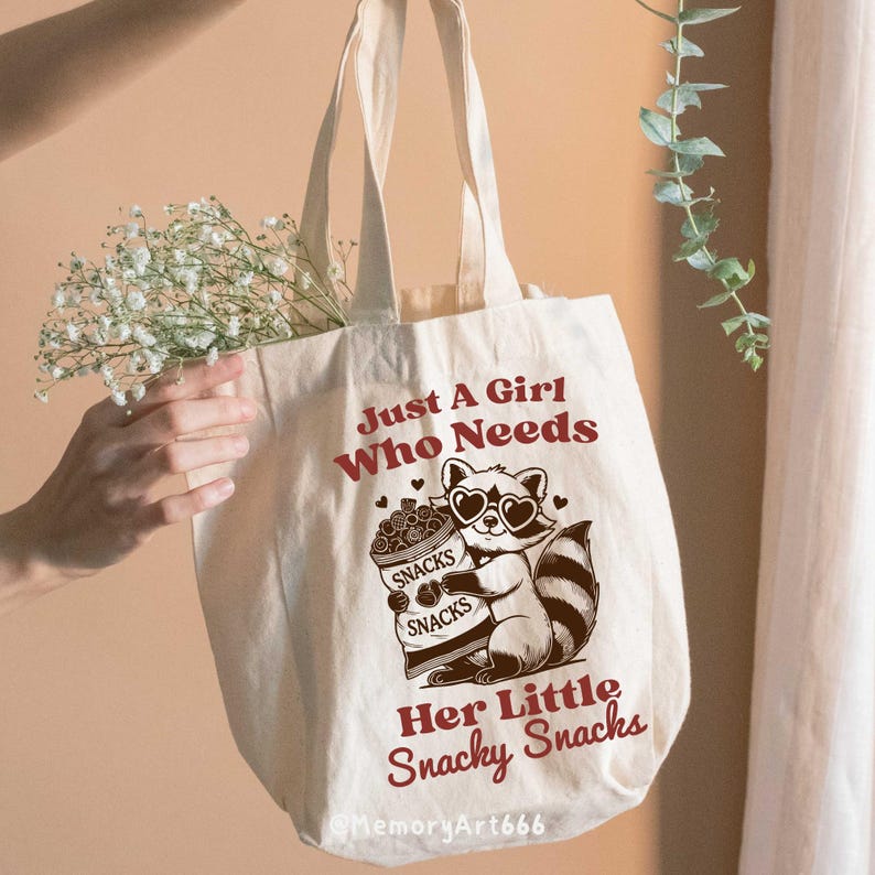 May include: A white canvas tote bag featuring a brown raccoon illustration holding a bag of snacks. The text on the bag reads "Just A Girl Who Needs Her Little Snacky Snacks".