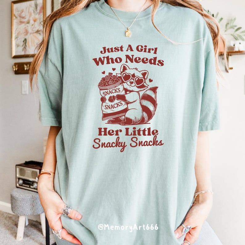 May include: A light blue t-shirt featuring a red graphic of a raccoon wearing heart-shaped sunglasses and holding a bag of snacks. The text "Just A Girl Who Needs Her Little Snacky Snacks" is printed on the shirt.