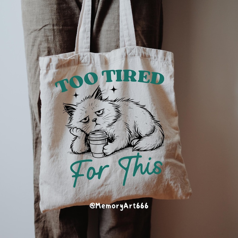 May include: Beige tote bag with the text "TOO TIRED For This" in teal. A grumpy cat illustration is featured below the text, holding a coffee cup. The bag has two handles and is made of a canvas-like material.