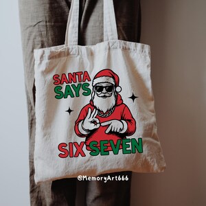 May include: Off-white canvas tote bag featuring a graphic of Santa Claus wearing sunglasses and a red suit. The text "SANTA SAYS SIX SEVEN" is printed in green and red. The bag has two black star accents.