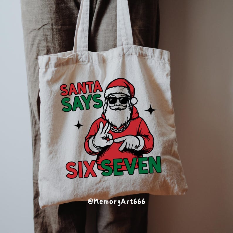May include: Off-white canvas tote bag featuring a graphic of Santa Claus wearing sunglasses and a red suit. The text "SANTA SAYS SIX SEVEN" is printed in green and red. The bag has two black star accents.