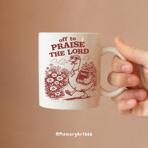 Op de afbeelding: Witte keramische mok met een illustratie van een gans met een hartvormige zonnebril en een kruis, die een bijbel draagt. De mok heeft de tekst "off to PRAISE THE LORD" in het rood.