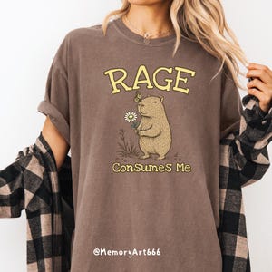 Funny capybara meme PNG SVG design featuring a cute vintage-style capybara holding a daisy flower with a butterfly resting on its head and bold retro “RAGE Consumes Me” typography. Funny sarcastic animal graphic with whimsical hand-drawn line art, soft neutral tones, and trendy unhinged aesthetic. Perfect for funny t-shirts, hoodies, mugs, tote bags, stickers, sublimation projects, Cricut SVG cut files, retro boutique apparel, instant digital download, 300 DPI transparent background.