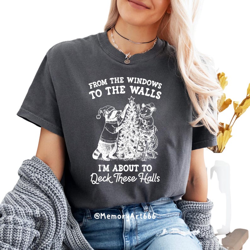 May include: Dark gray t-shirt with a white graphic featuring a raccoon and a mouse decorating a Christmas tree. The text reads "From the windows to the walls, I'm about to Deck These Halls."