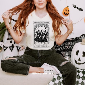 May include: Cream-colored t-shirt featuring a black and white tarot card design with the text "THE WITCH." The graphic depicts three witches dancing under a crescent moon. The shirt is paired with black jeans and white sneakers, creating a casual, witchy aesthetic.