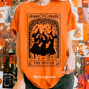 May include: Orange t-shirt featuring a black graphic of three witches dancing, framed by roses, a crescent moon, and stars. The text "THE WITCH" is below the image. The shirt also has the text "VIII" at the top.