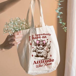 Op de afbeelding: Een witte canvas tote bag met een roze en bruine illustratie van een Highland koe die een zonnebril en een roze strik draagt. De tekst "Apparently I have an Attitude Who Knew" is in rood op de tas gedrukt.