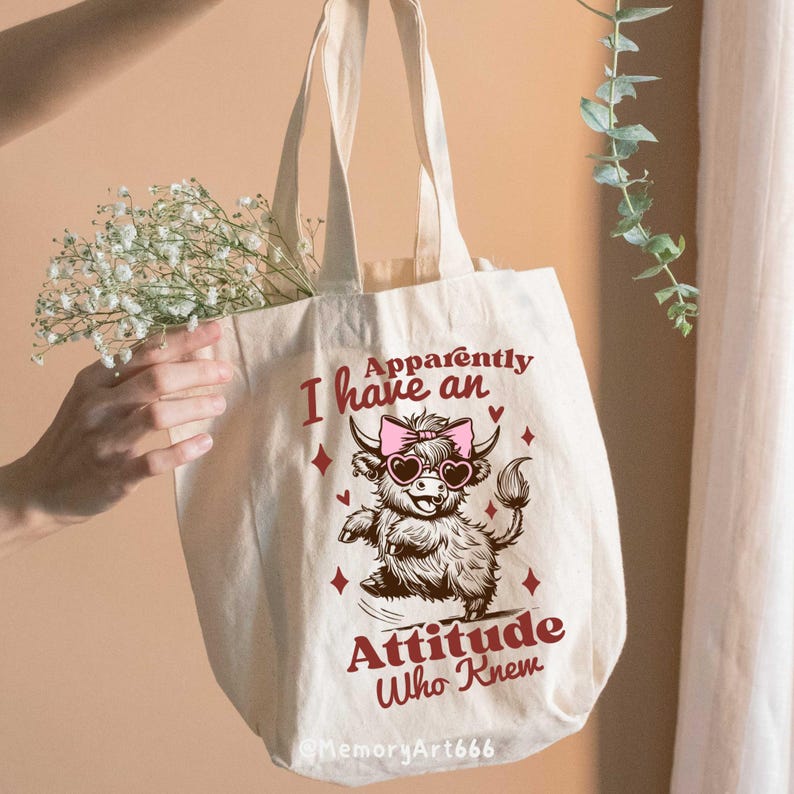 Op de afbeelding: Een witte canvas tote bag met een roze en bruine illustratie van een Highland koe die een zonnebril en een roze strik draagt. De tekst "Apparently I have an Attitude Who Knew" is in rood op de tas gedrukt.