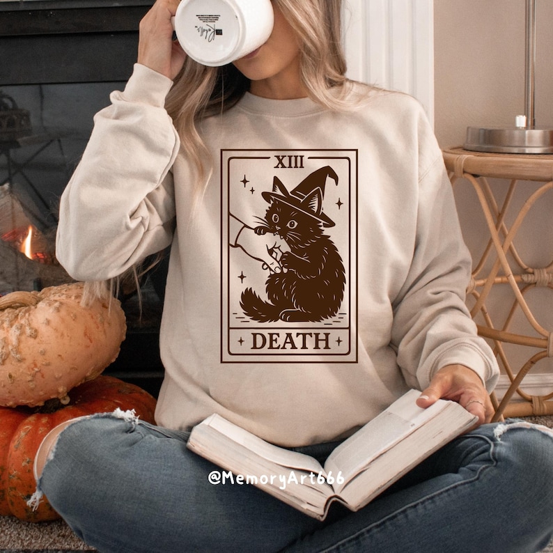 May include: Beige sweatshirt featuring a tarot card design with a black cat wearing a witch hat. The card reads "XIII DEATH" in a rectangular frame. The sweatshirt is worn by a person holding a mug and a book.