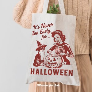 Puede incluir: Una bolsa de tela blanca con un gr&aacute;fico rojo de una mujer esqueleto tallando una calabaza con un gato que lleva un sombrero de bruja. El texto en la bolsa dice "It's Never Too Early for... HALLOWEEN @MemoryArt666"
