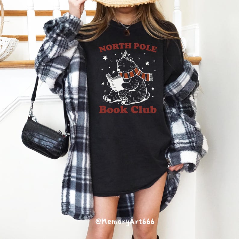 May include: Black t-shirt with a white graphic of a bear wearing a scarf and reading a book. The text "North Pole Book Club" is printed in red on the shirt.