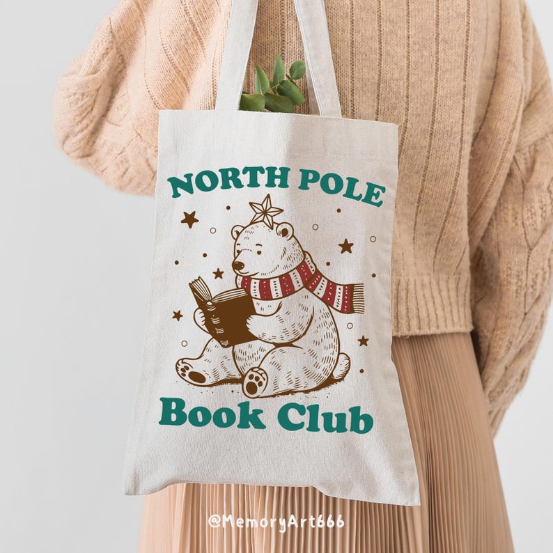 May include: A white canvas tote bag with a brown illustration of a polar bear wearing a red and white striped scarf and reading a book. The text "North Pole Book Club" is printed in green on the bag.