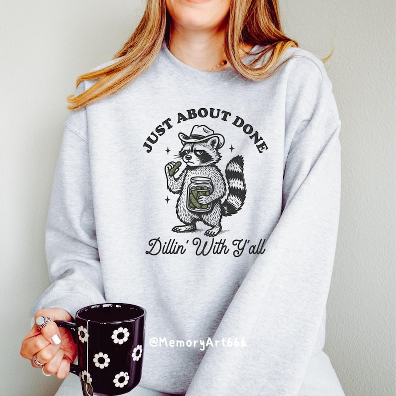 K&ouml;nnte beinhalten: Hellgraues Sweatshirt mit einer Grafik eines Waschb&auml;ren mit Cowboyhut, der ein Einmachglas h&auml;lt, und dem Text "JUST ABOUT DONE Dillin' With Y'all". Eine schwarze Tasse mit wei&szlig;en Blumenmustern wird in der Hand gehalten.