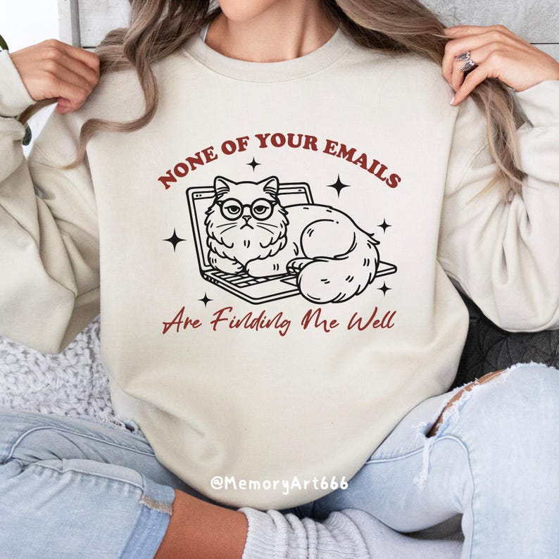 May include: A beige sweatshirt with a graphic of a cat wearing glasses and sitting on a laptop. The text on the sweatshirt reads "None of your emails are finding me well" and "@MemoryArt666".