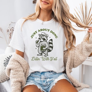 K&ouml;nnte beinhalten: Wei&szlig;es T-Shirt mit einer Illustration eines Waschb&auml;ren mit Hut, der ein Glas h&auml;lt. Der Text lautet "JUST ABOUT DONE" dar&uuml;ber und "Dillin' With Y'all" darunter. Das T-Shirt wird von einer Person getragen.