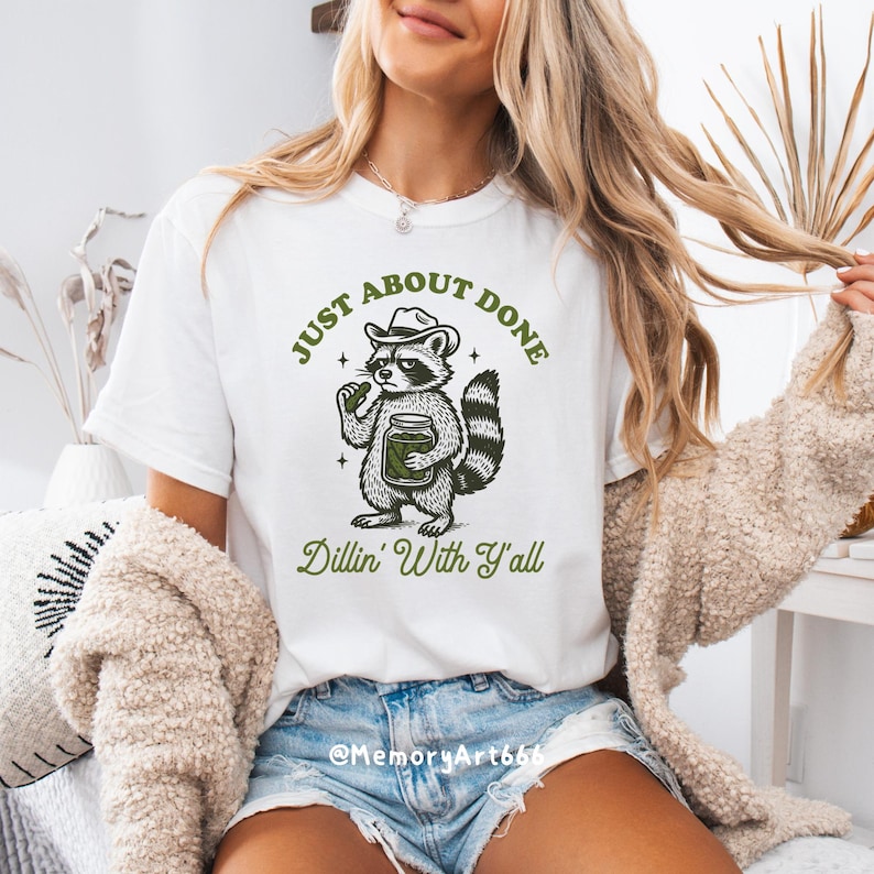 K&ouml;nnte beinhalten: Wei&szlig;es T-Shirt mit einer Illustration eines Waschb&auml;ren mit Hut, der ein Glas h&auml;lt. Der Text lautet "JUST ABOUT DONE" dar&uuml;ber und "Dillin' With Y'all" darunter. Das T-Shirt wird von einer Person getragen.