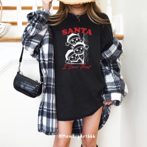 May include: Black t-shirt featuring a graphic of three cats wearing Santa hats peeking around a corner. The text "SANTA I Saw That" is printed below the cats.