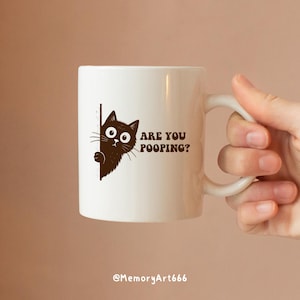 May include: White ceramic mug with a brown cat graphic and the text "ARE YOU POOPING?". The mug has a curved handle and is held by a hand. The bottom of the mug has the text "@MemoryArt666".