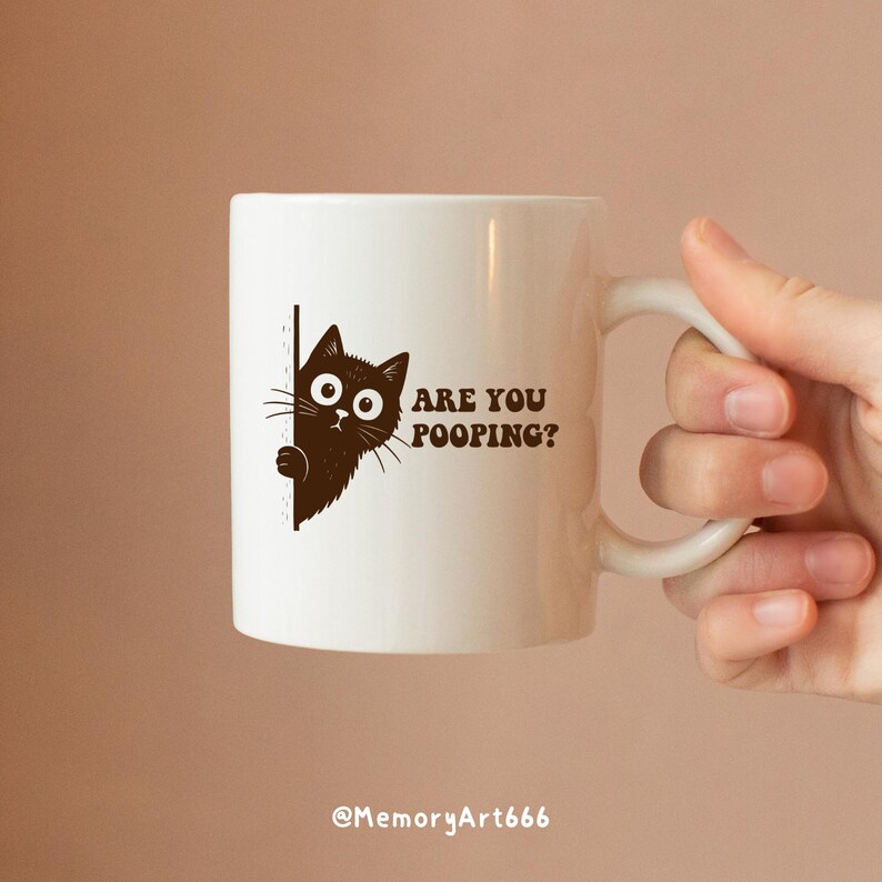 May include: White ceramic mug with a brown cat graphic and the text "ARE YOU POOPING?". The mug has a curved handle and is held by a hand. The bottom of the mug has the text "@MemoryArt666".