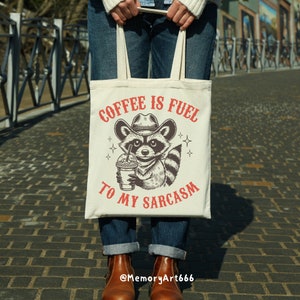 Puede incluir: Una bolsa de tela blanca con una ilustraci&oacute;n en blanco y negro de un mapache con un sombrero de vaquero y sosteniendo una taza de caf&eacute;. El texto "Coffee is fuel to my sarcasm" est&aacute; impreso en rojo en la bolsa.
