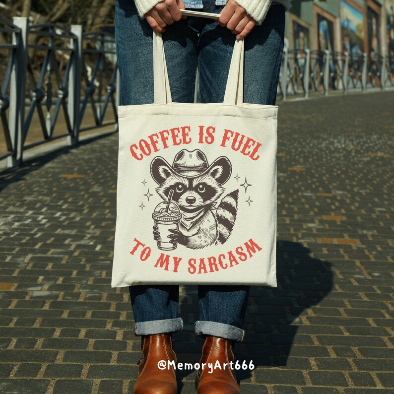Puede incluir: Una bolsa de tela blanca con una ilustraci&oacute;n en blanco y negro de un mapache con un sombrero de vaquero y sosteniendo una taza de caf&eacute;. El texto "Coffee is fuel to my sarcasm" est&aacute; impreso en rojo en la bolsa.