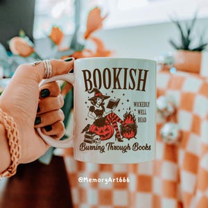 May include: White ceramic mug with a witch-themed design. The mug features the word "BOOKISH" above an illustration of a witch reading a book, with the text "WICKEDLY WELL READ" and "Burning Through Books".