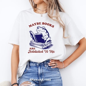 May include: White t-shirt featuring a frog reading a book, with the text "Maybe Books Are Addicted To Me" in maroon. The frog illustration is in blue and black. The shirt is a casual wear item.