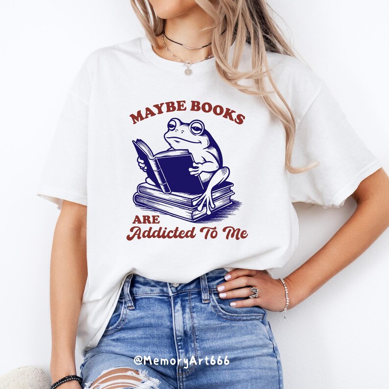 May include: White t-shirt featuring a frog reading a book, with the text "Maybe Books Are Addicted To Me" in maroon. The frog illustration is in blue and black. The shirt is a casual wear item.