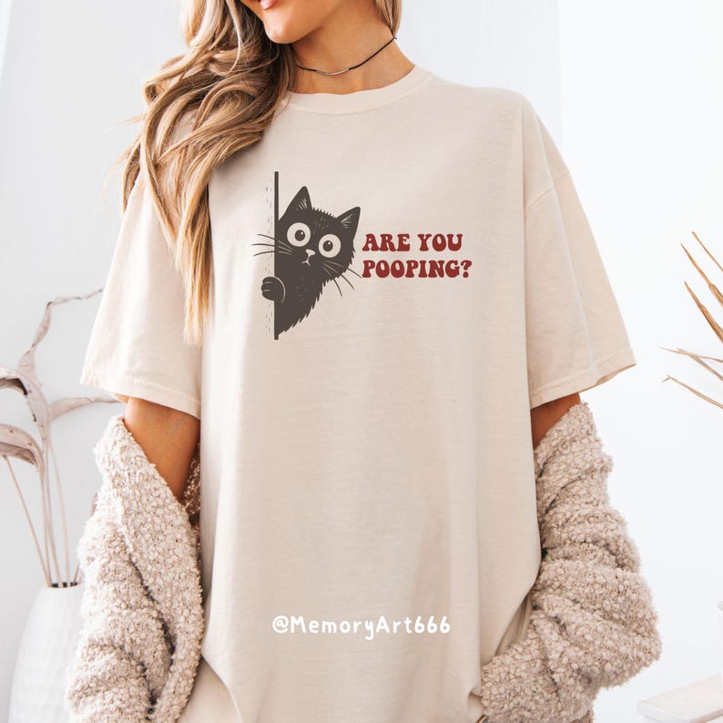 May include: Beige t-shirt with a black cat graphic peeking from behind a line, with the text "ARE YOU POOPING?" in red. The shirt has short sleeves and a relaxed fit.