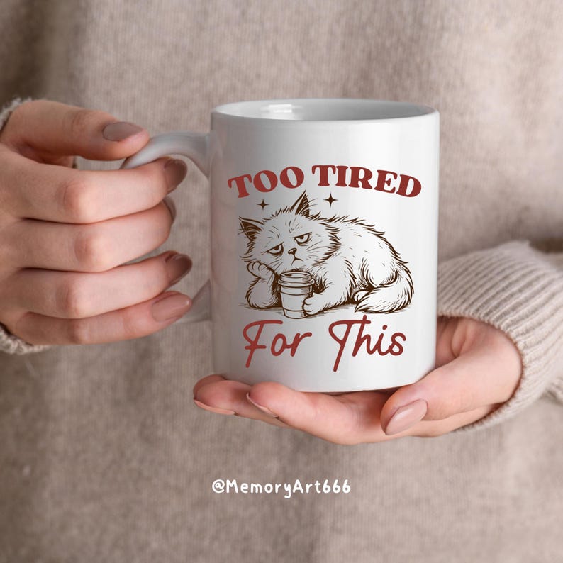 May include: White ceramic mug featuring a grumpy cat illustration with the text "TOO TIRED For This" in red. The cat is drawn in a relaxed pose, holding a coffee cup. The mug has a curved handle.