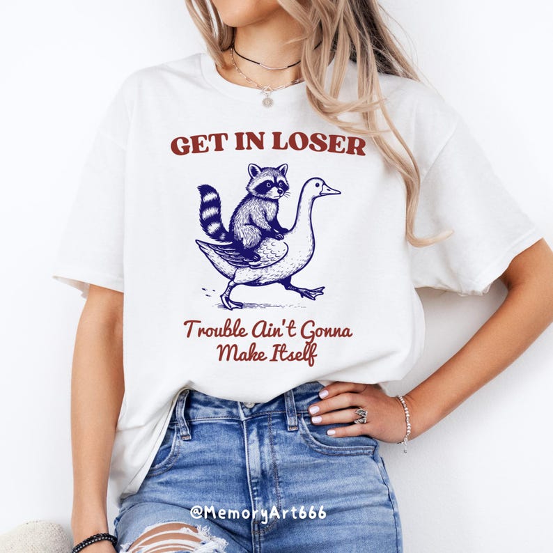 May include: White t-shirt with a blue and black illustration of a raccoon riding a goose. The text "Get in Loser, Trouble Ain't Gonna Make Itself" is printed in red on the shirt.