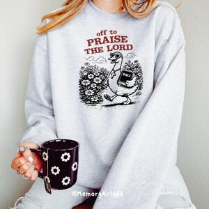 Op de afbeelding: Een lichtgrijs sweatshirt met de tekst "off to PRAISE THE LORD" in bordeauxrood. De afbeelding toont een cartoon gans met een zonnebril en een kruis, die een bijbel draagt. De gans loopt door een bloemenveld.